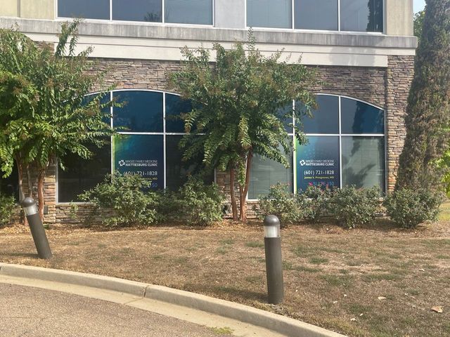Exterior of an office building with signage on two glass windows, framed by small trees, shrubs, and a curb in front.