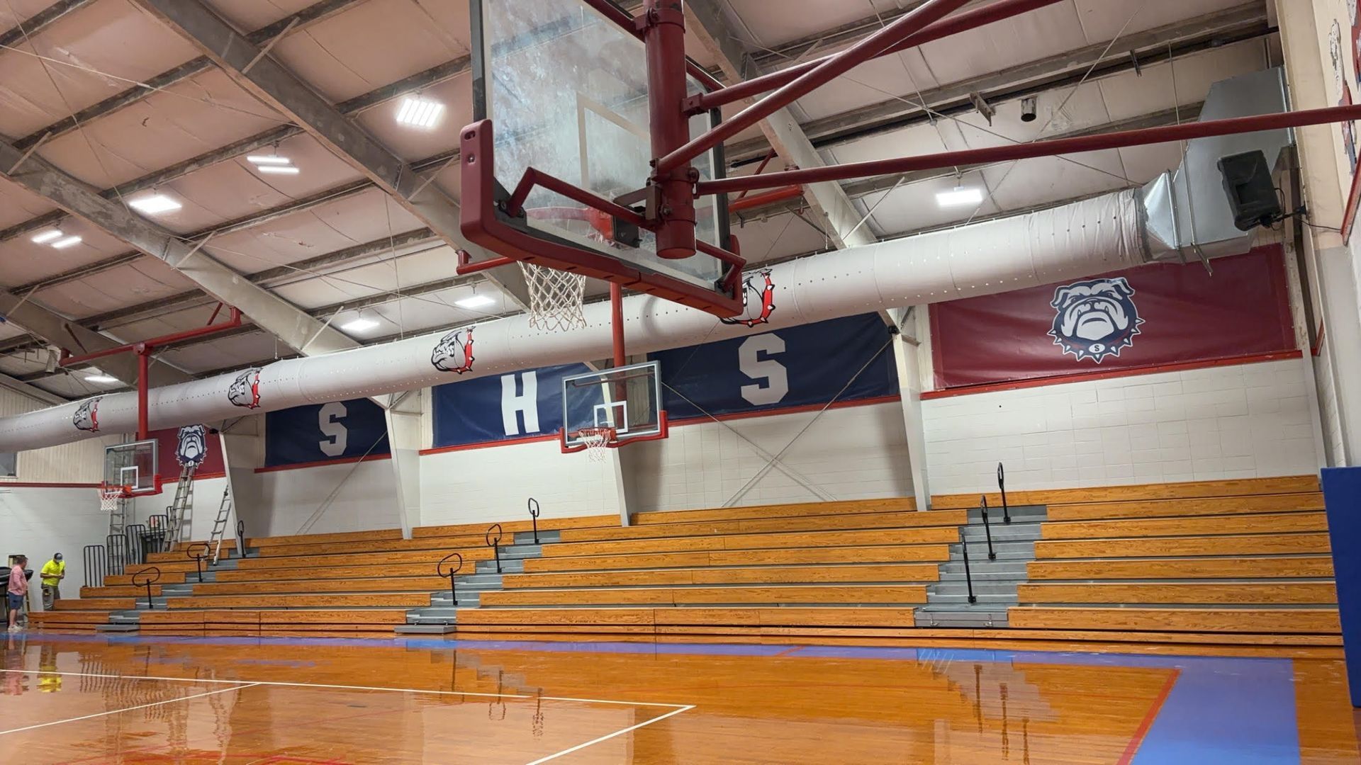 Interior view of a gymnasium featuring wood bleachers, a basketball hoop, and blue banners with the letter 