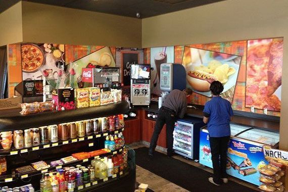 An indoor convenience store features stocked shelves in the foreground and a food preparation area with two staff members.