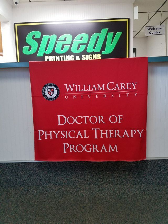 A red banner for the William Carey University Doctor of Physical Therapy program hanging in a Speedy Printing & Signs shop.