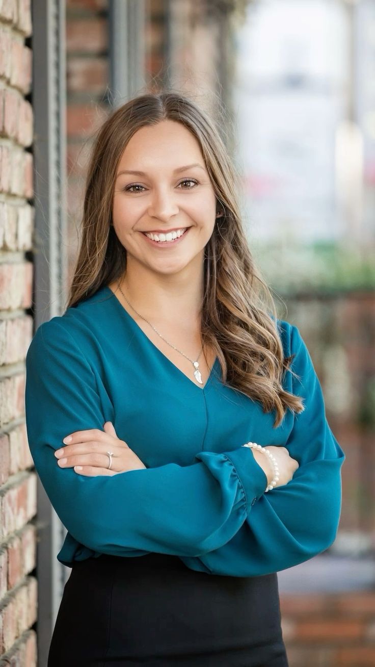 Woman with crossed arms smiles, wearing teal blouse and black skirt, standing by a brick wall.