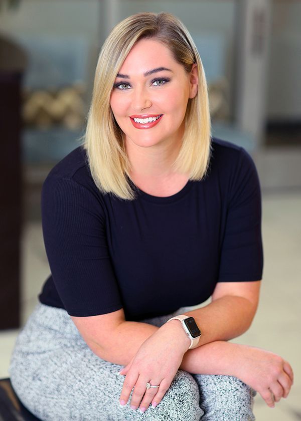 Woman with blonde hair, smiling in a dark top and patterned skirt, sitting indoors.