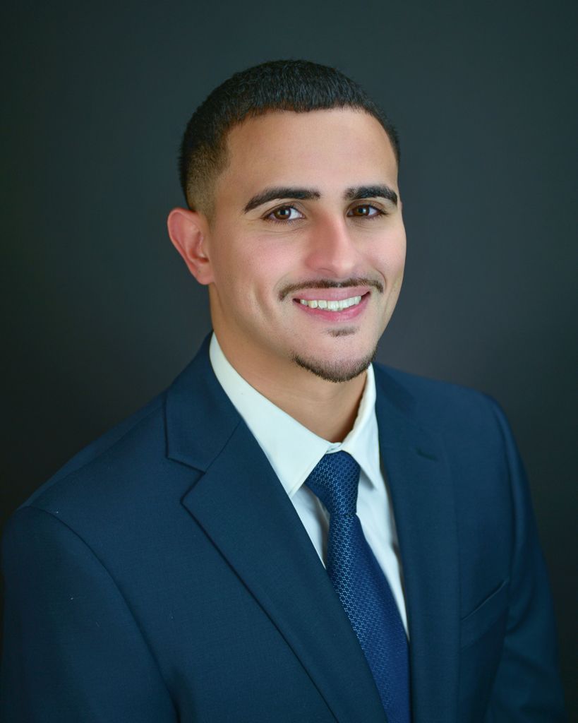 Man in navy suit, smiling, dark hair, blue tie, against dark gray background.
