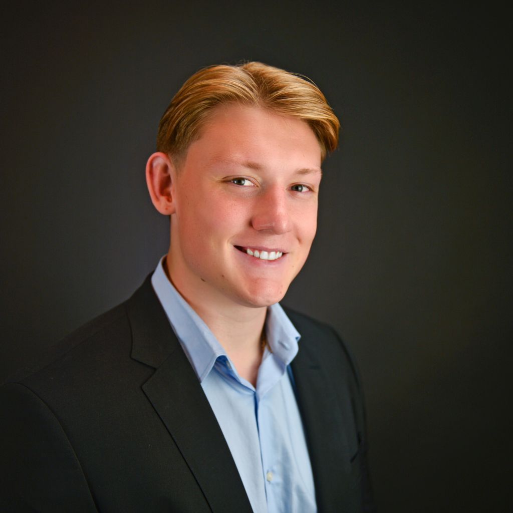 Man in a blue shirt and black blazer smiling against a dark gray background.