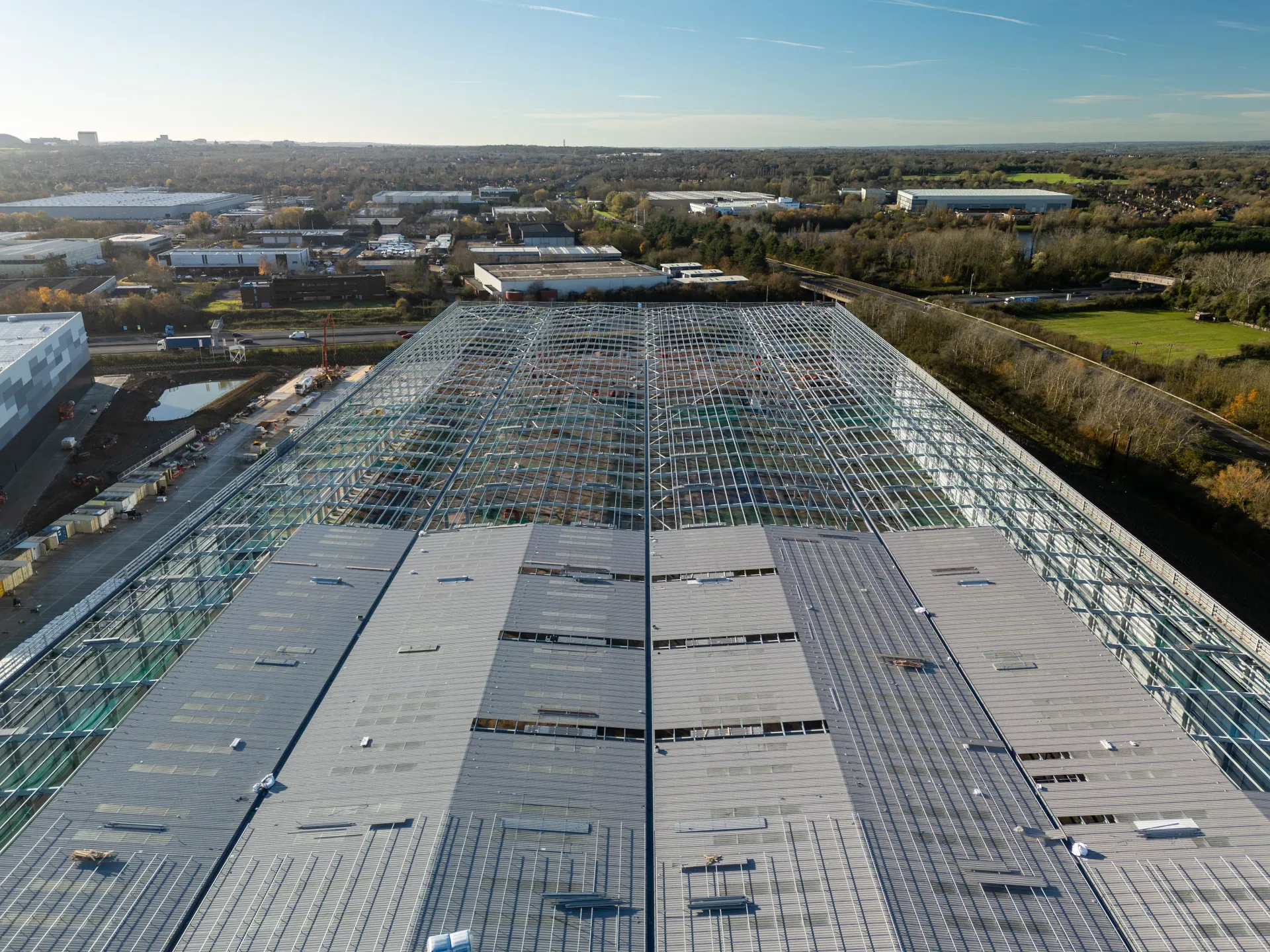 Large warehouse under construction. Steel framework, partially completed roof, and surrounding industrial area are visible.