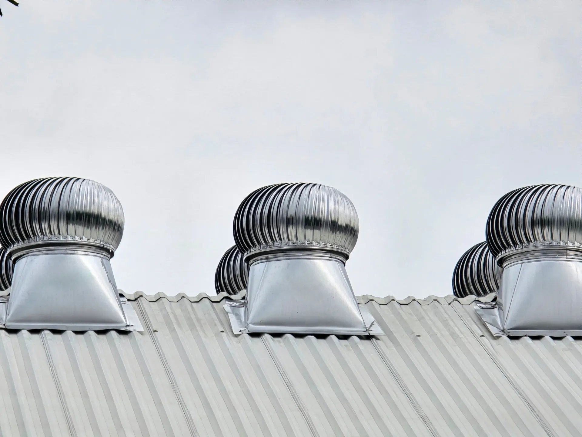 Metallic rooftop turbine vents on a corrugated roof against a cloudy sky.