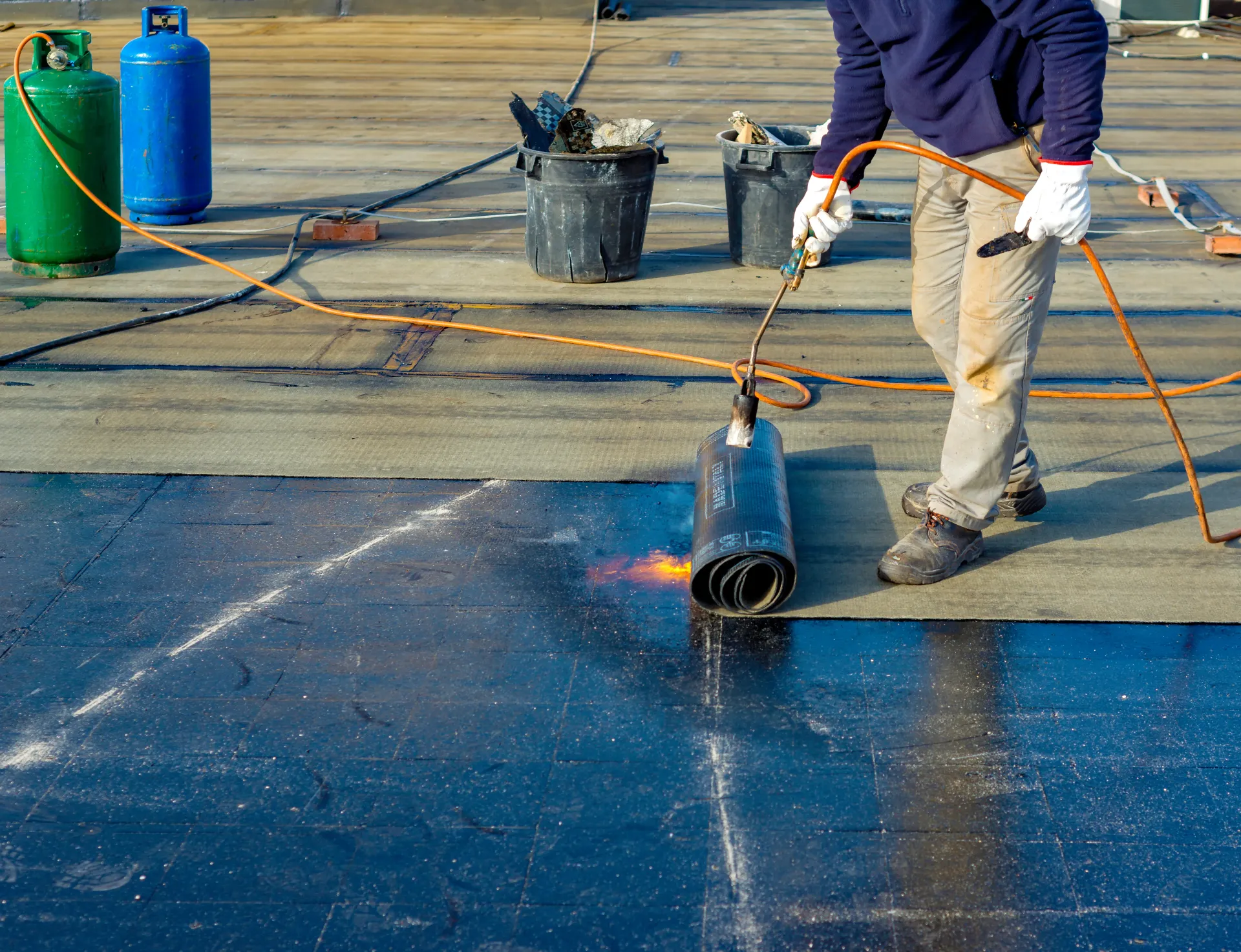 Roofer using a torch to install a dark roofing membrane on a flat roof. Propane tanks nearby.