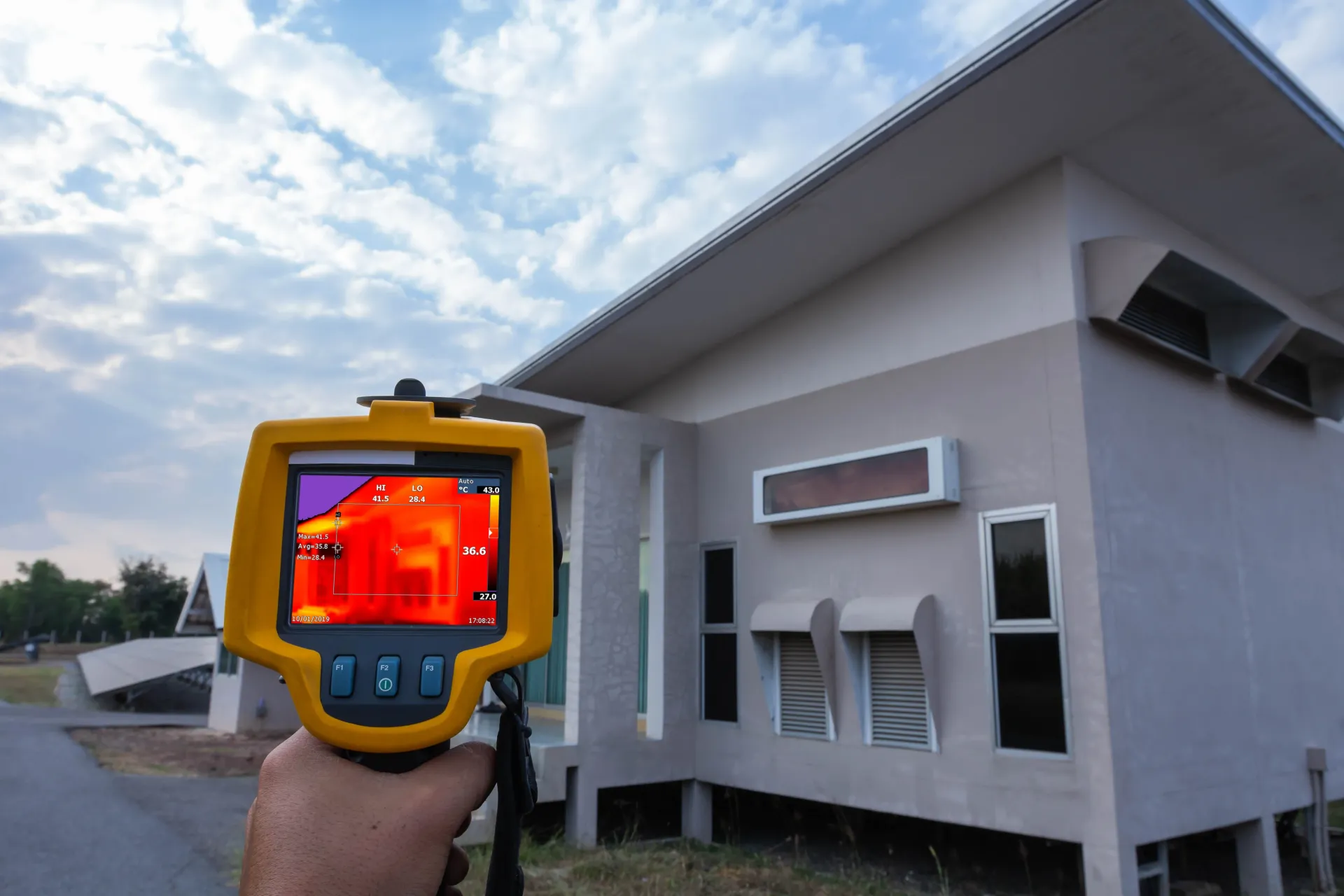 A person uses a thermal imaging camera to inspect a light-colored building under a cloudy sky.