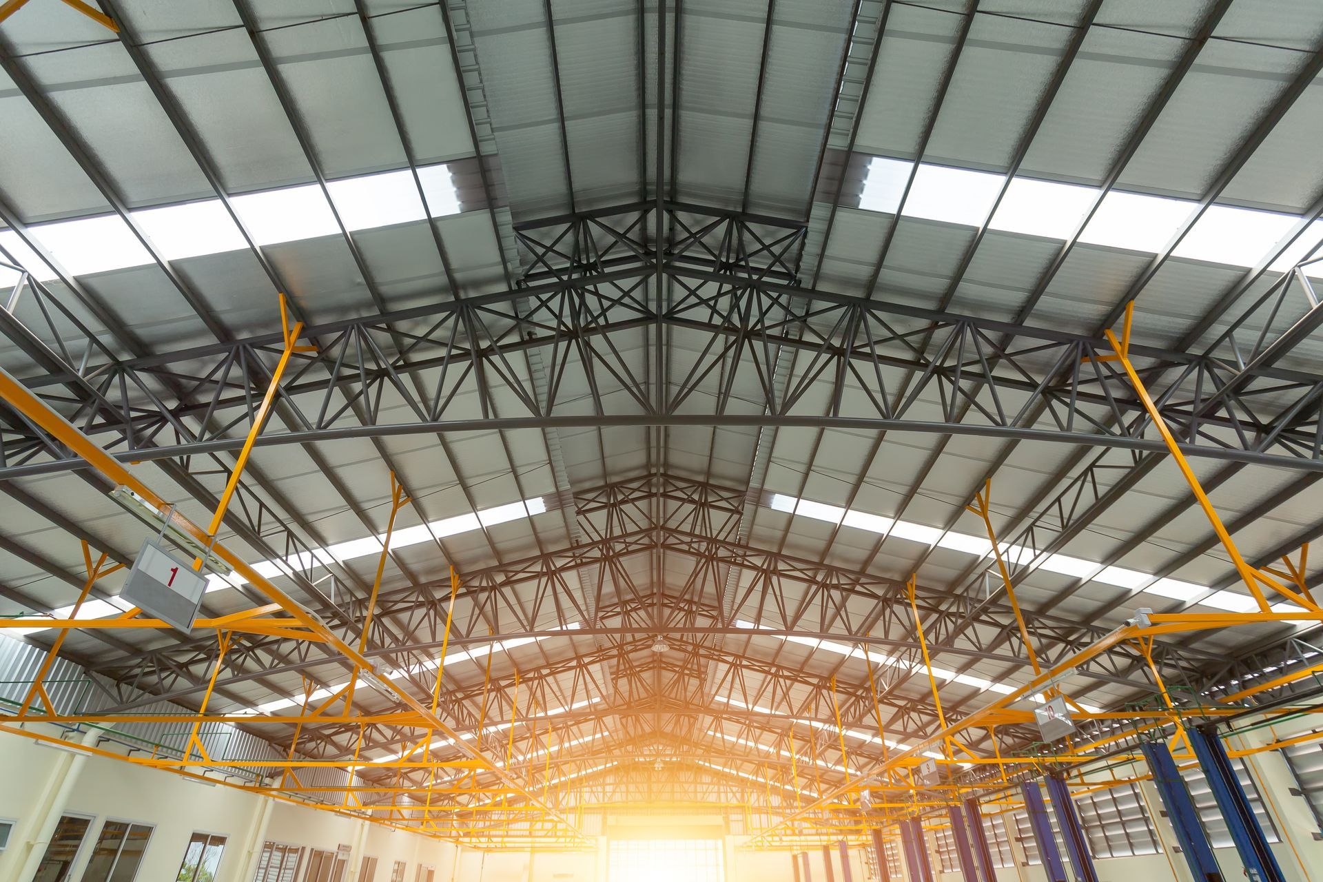 Warehouse interior with a metal truss roof and skylights, lit by the sun.