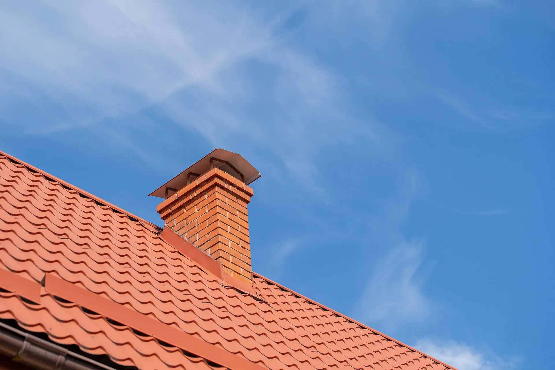 Red tiled roof with a brick chimney against a bright blue sky with wispy clouds.