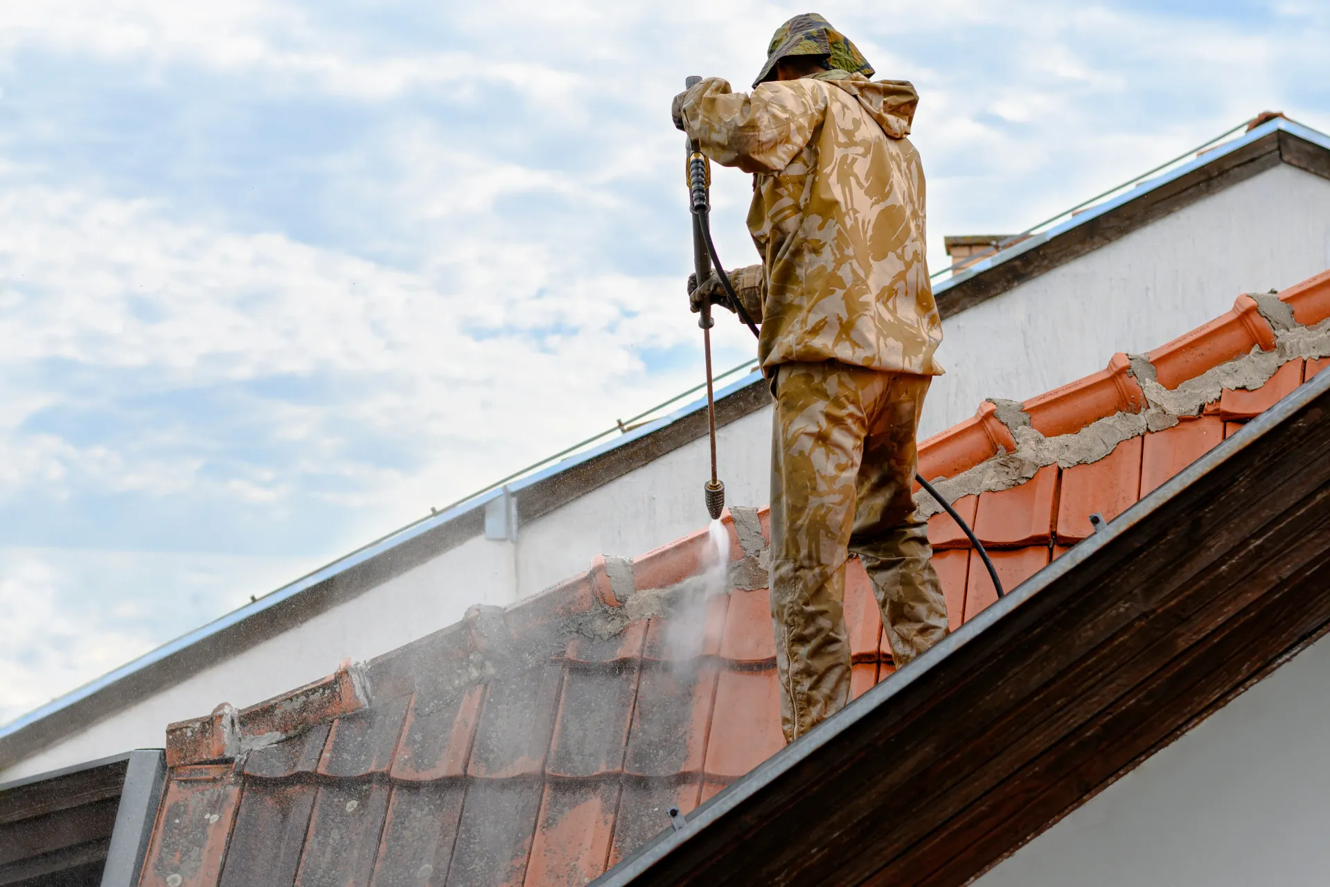 Person in protective gear pressure washes a brick wall on a rooftop; water sprays everywhere.