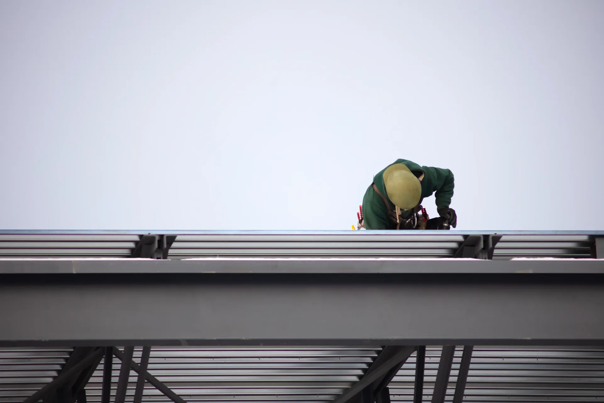 Worker in green suit and yellow helmet on metal structure, against a cloudy sky.