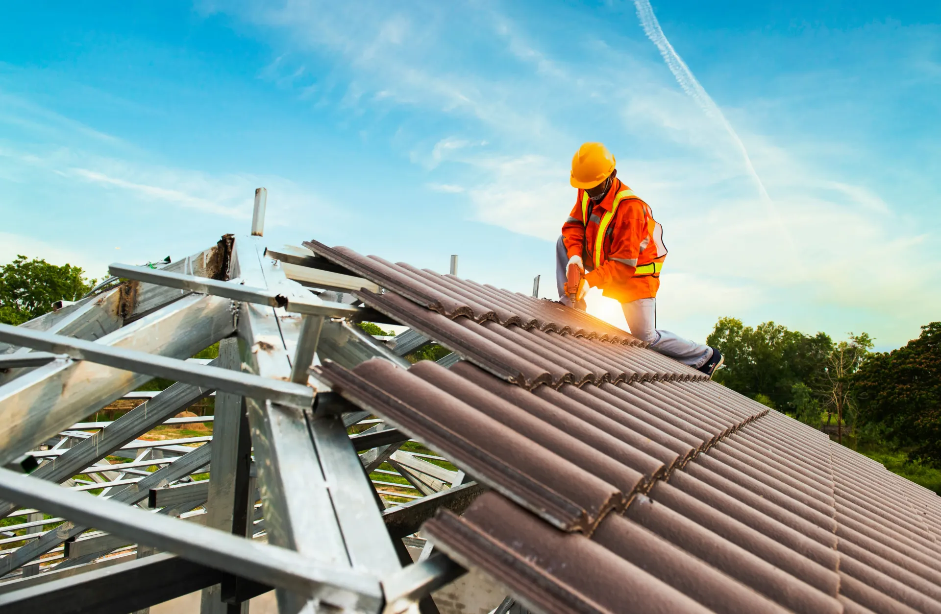 Roofer in orange vest and hard hat installing brown tiles on a metal-framed roof under a blue sky.