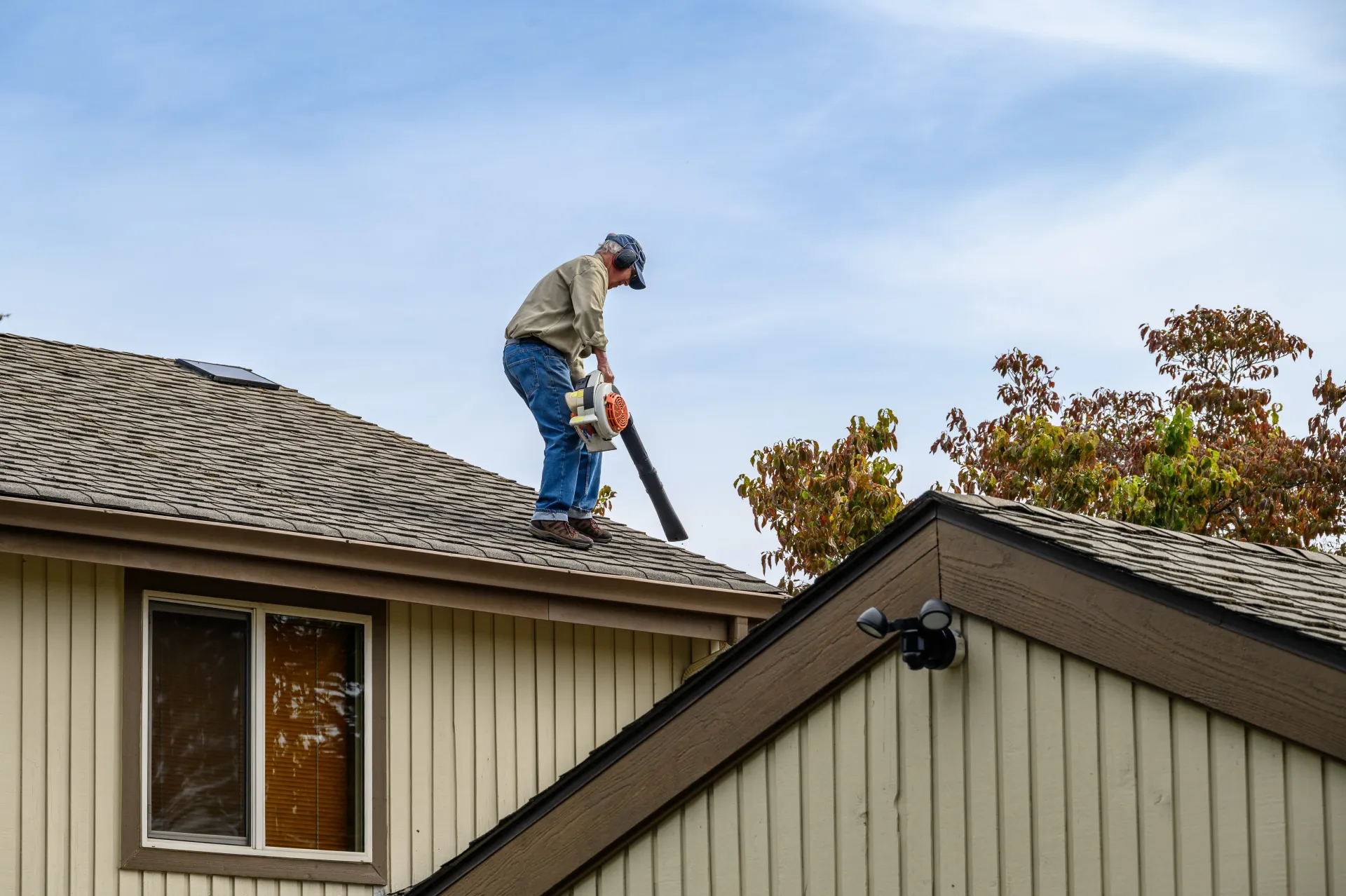 Man using a leaf blower on a rooftop, clearing debris. Blue sky, neutral-colored house.