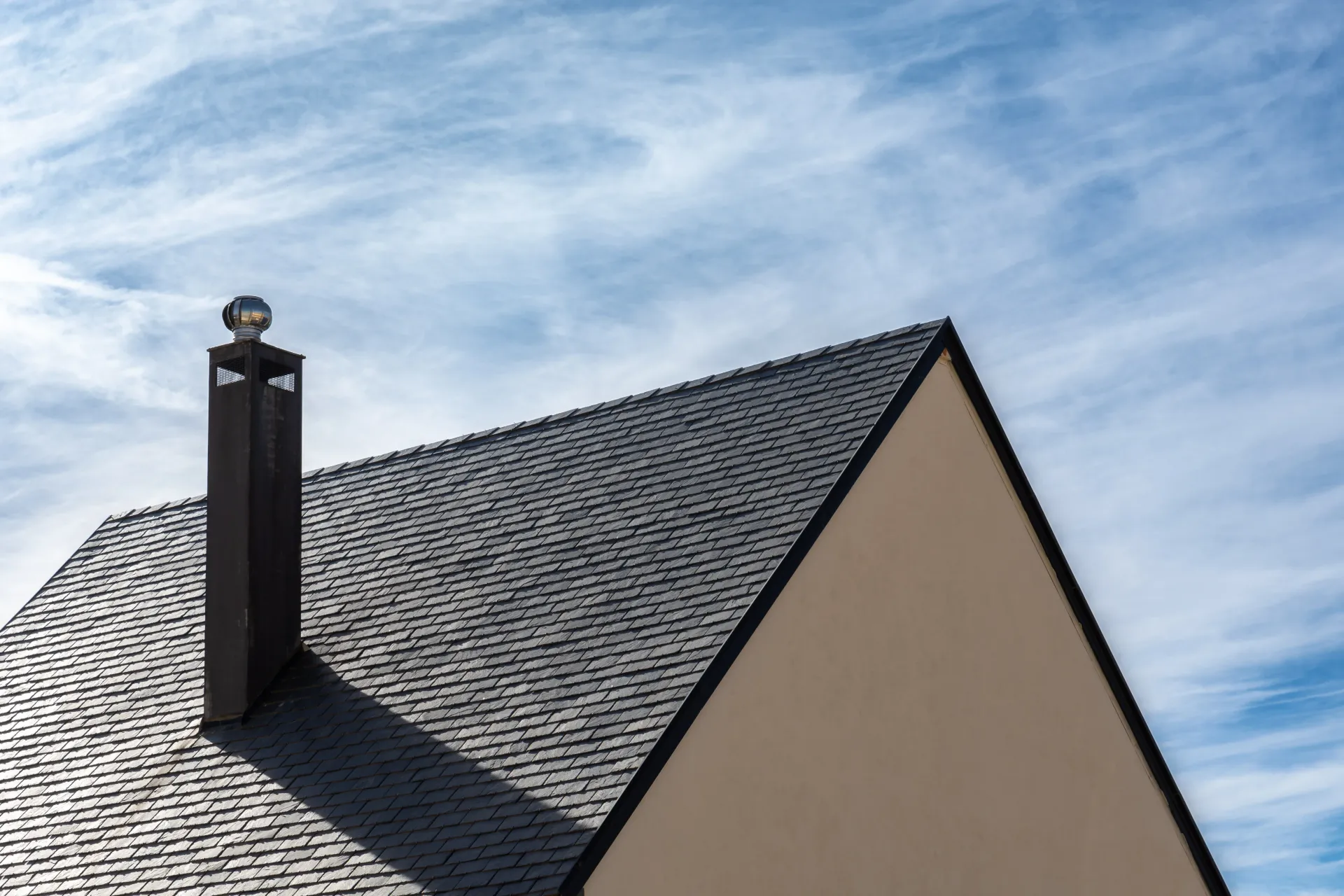 Roof of a house with dark shingles, black chimney, against a blue sky with streaky clouds.