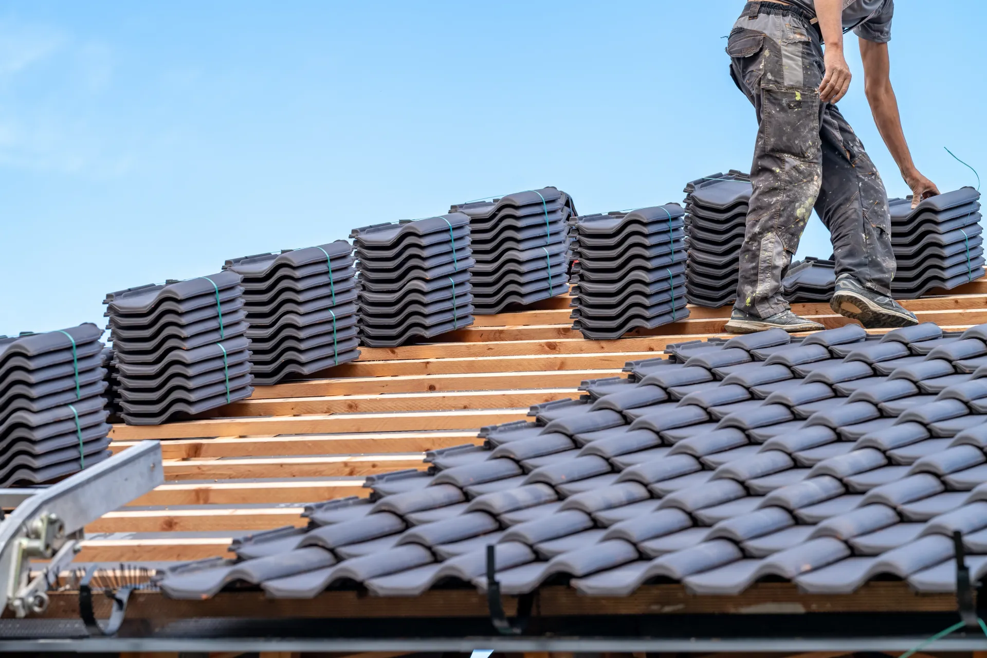 Roofer laying dark gray tiles on a wooden roof framework under a blue sky.