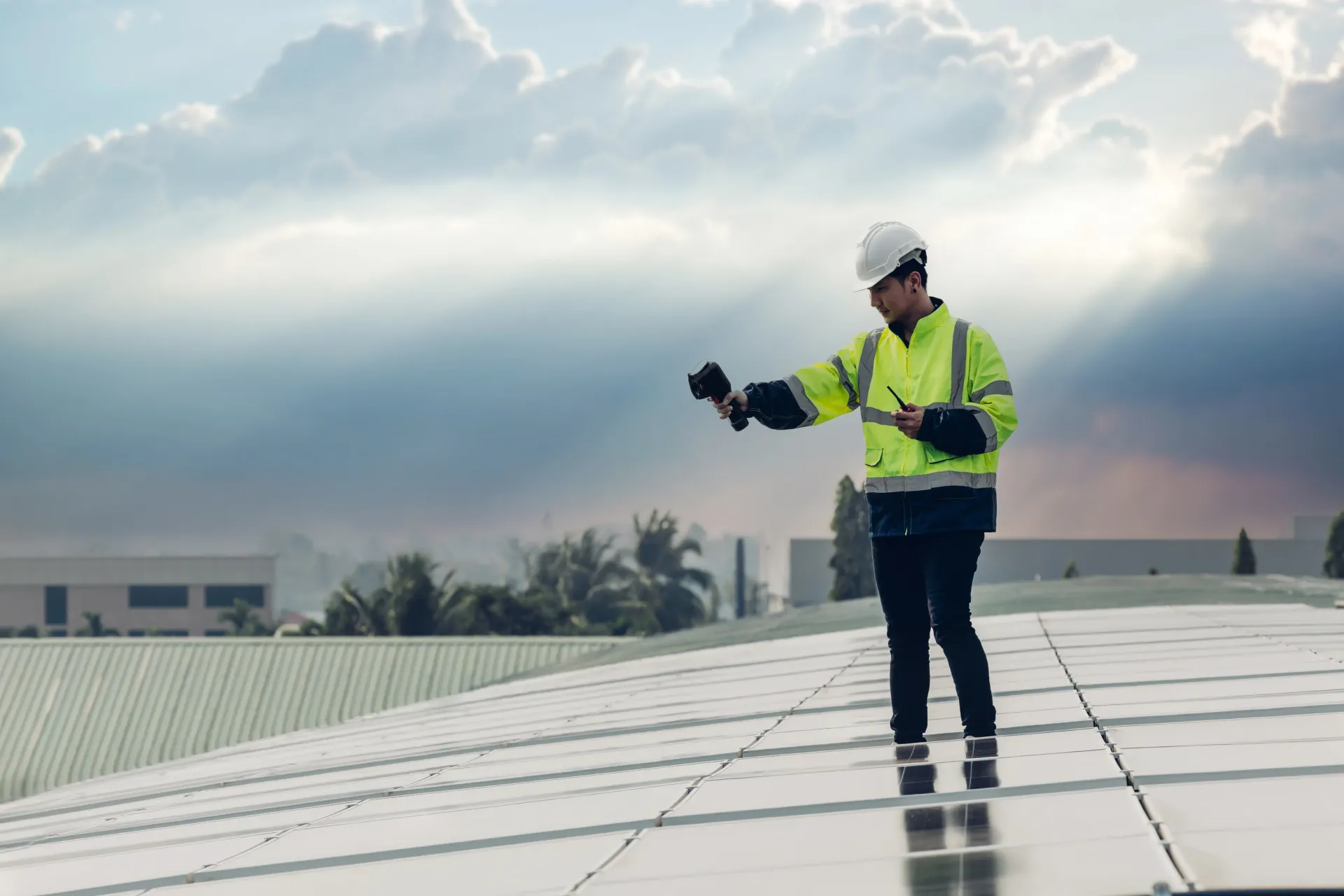 Worker in safety gear inspecting solar panels on a rooftop under a cloudy sky.