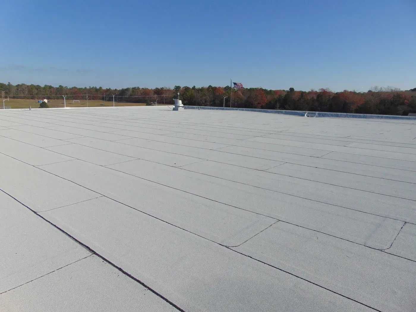 Flat white roof with visible seams, under a clear blue sky, with a treeline in the distance.
