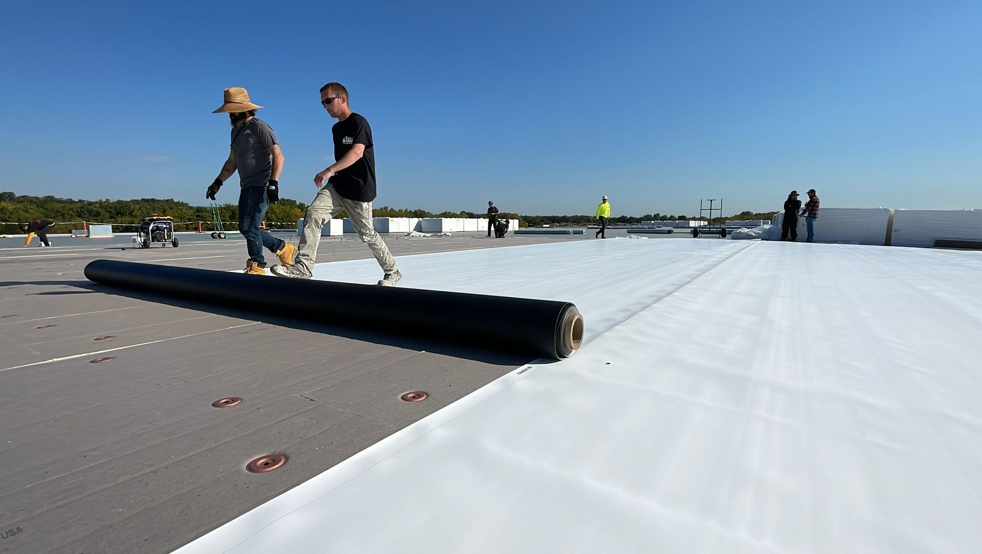 Workers rolling out roofing material on a flat rooftop. Bright blue sky overhead.