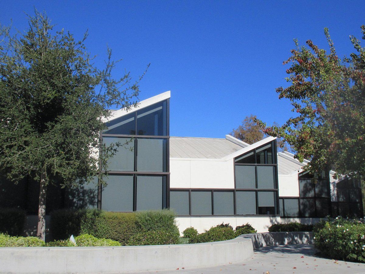 White modern building with angled glass windows and a blue sky behind trees.