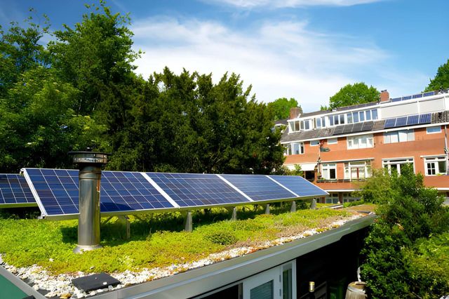 Solar panels on a green roof with a residential building in the background. Blue sky with trees.