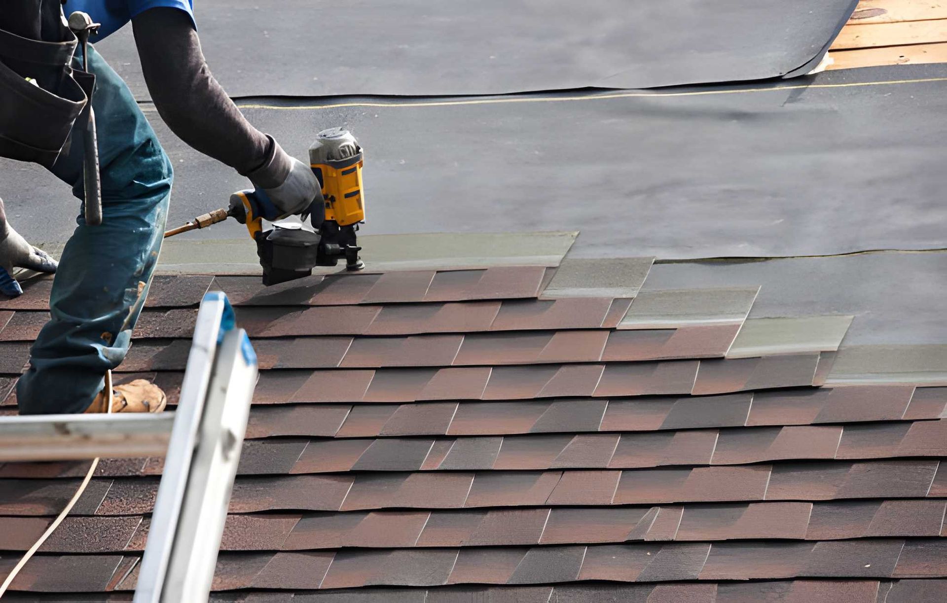 Roofer using a nail gun to install asphalt shingles on a roof. Brown shingles and dark underlayment are visible.