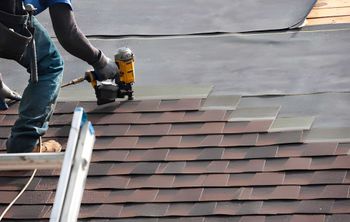 Roofer using a nail gun to install asphalt shingles on a roof. Brown shingles and dark underlayment are visible.