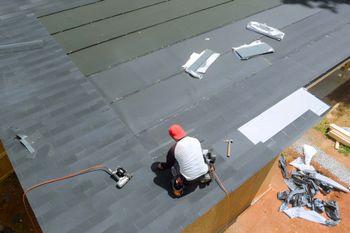 Roofer installing dark gray shingles on a roof. Person wearing a red cap and white shirt, using tools.