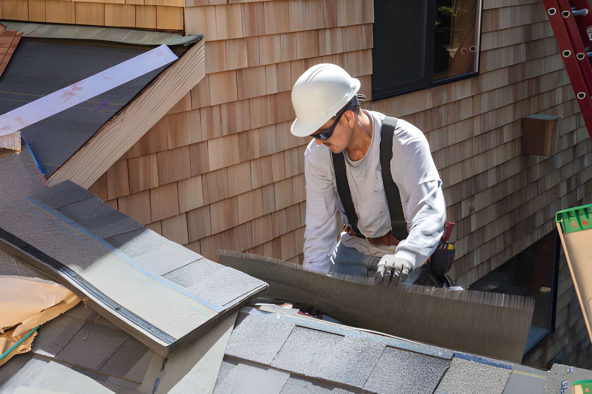 Roofer in white hard hat and gloves working on a roof with gray shingles and wooden siding.