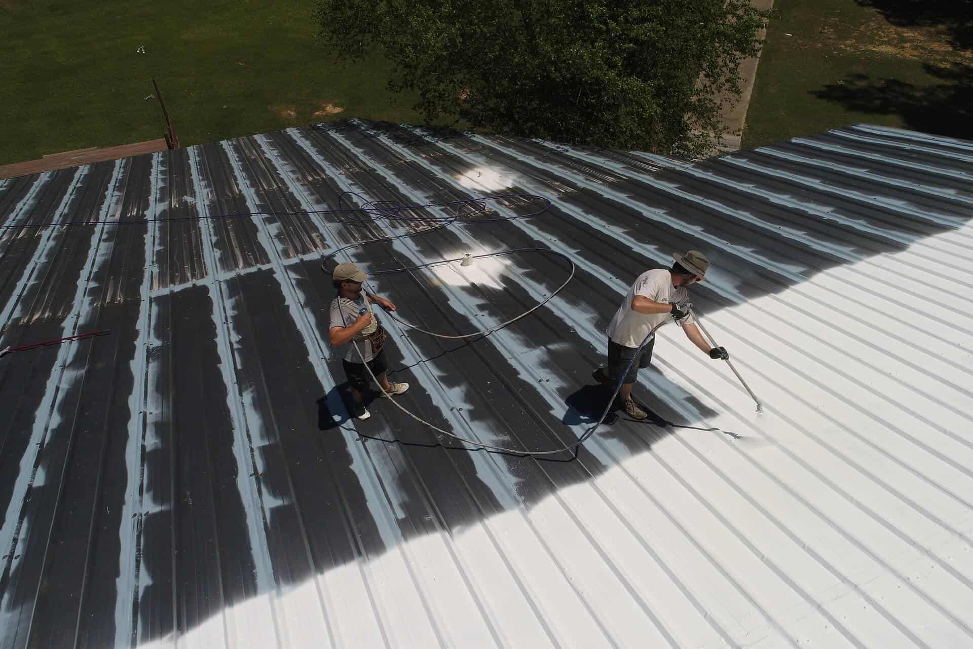 Two people spray painting a metal roof white on a sunny day.