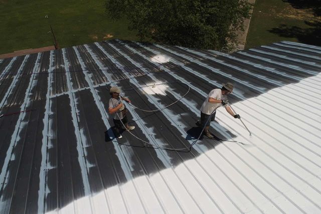 Two people spray painting a metal roof white on a sunny day.