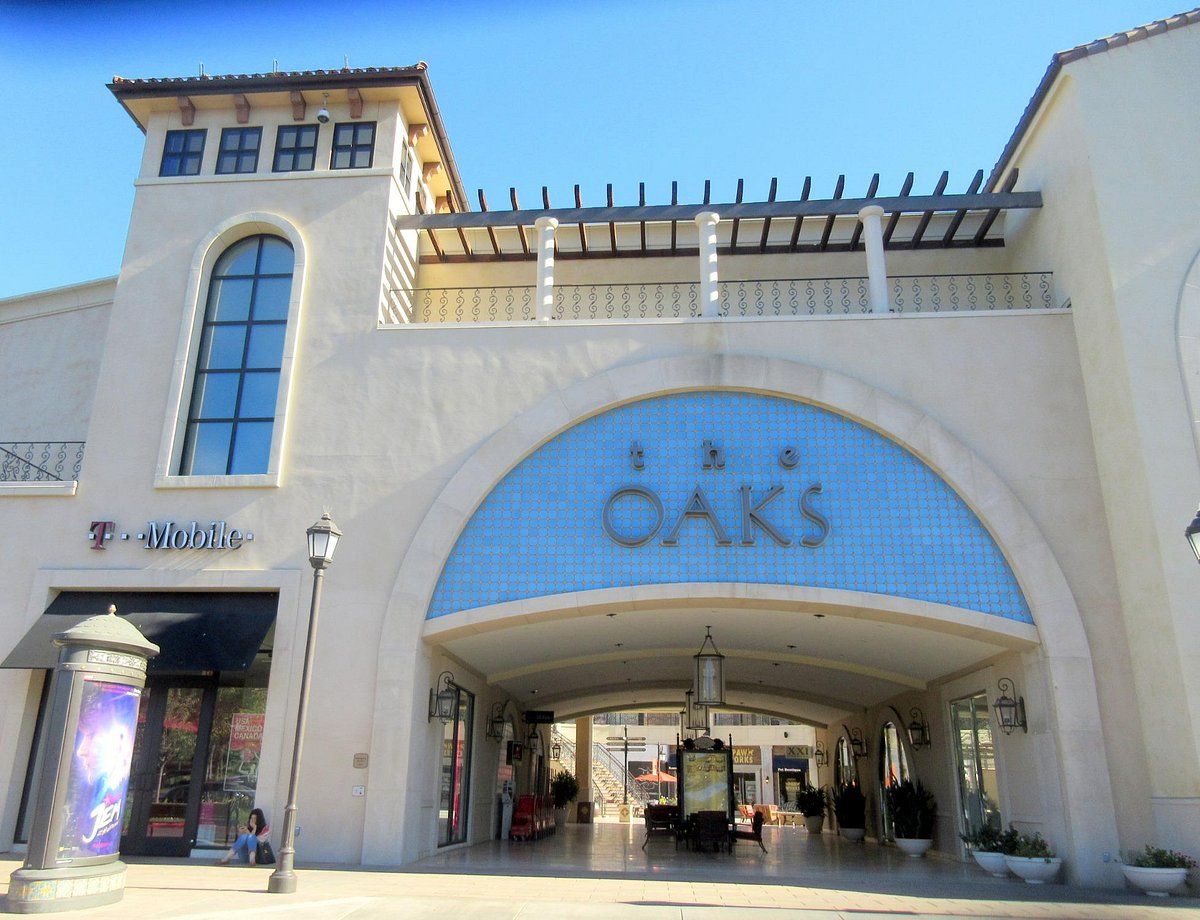 Exterior of The Oaks mall with arched entrance and T-Mobile store. Blue sky, neutral tones.