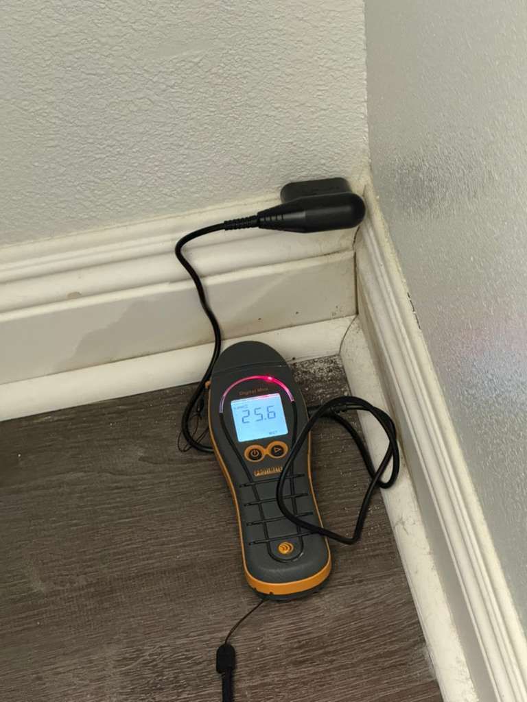 A handheld moisture meter resting on a floor corner, reading 25.8% moisture near a wall and baseboard.