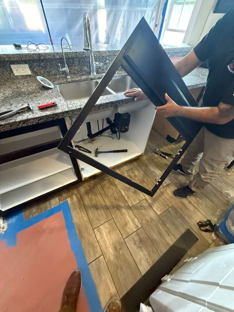 A worker holds a dark, rectangular cabinet frame in front of an open kitchen sink cabinet during installation.