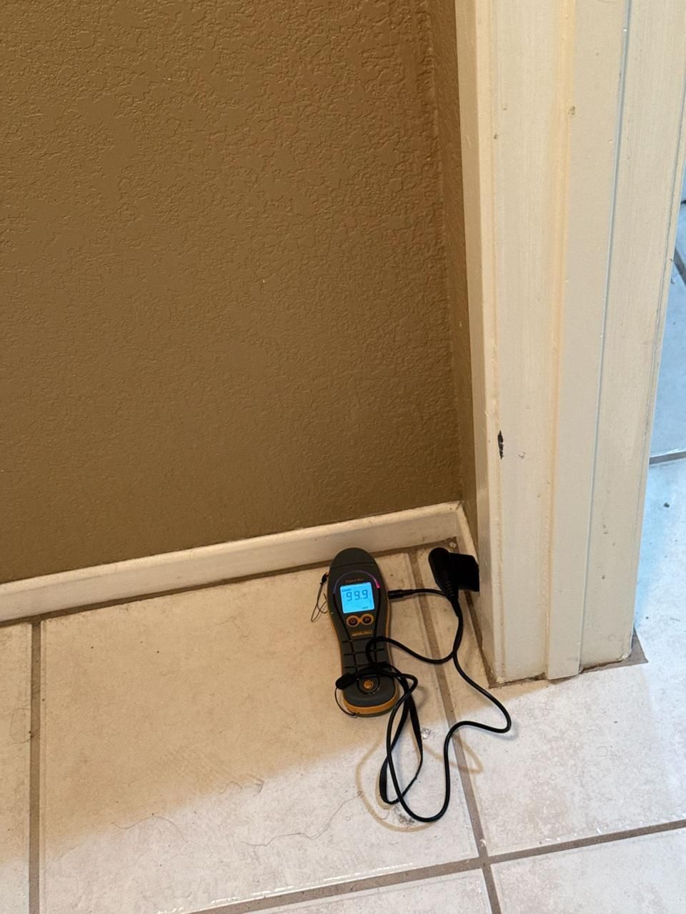 A moisture meter with a digital display rests on a tiled floor against a beige wall near a door frame.
