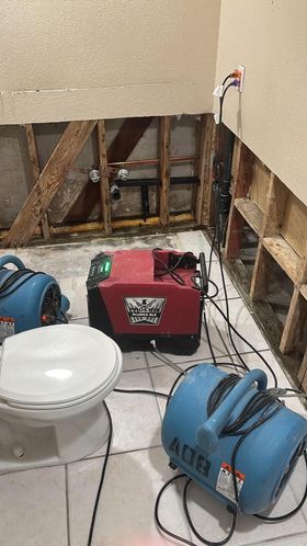 A bathroom under repair with wall panels removed, showing exposed studs and plumbing, with drying fans and a dehumidifier.