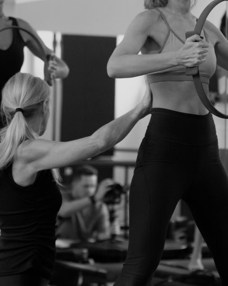 A yoga instructor assists a student in a lunge position with arms raised in a sunlit studio.