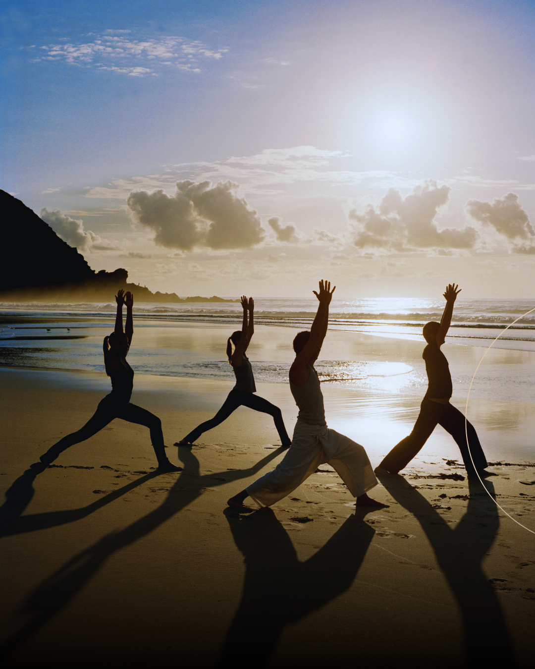 Four people in silhouette perform yoga poses on a sandy beach at sunrise, their reflections visible on the wet shore.