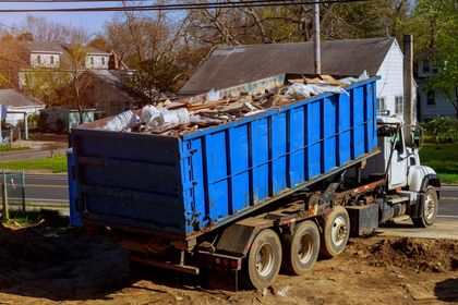 A dumpster truck is driving down a dirt road next to a house.