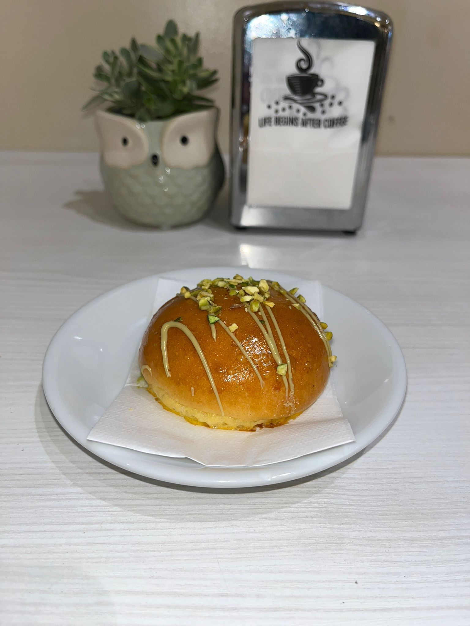 Un dolce glassato con pistacchi su un piatto bianco, una piccola pianta grassa a forma di gufo e un porta tovaglioli.
