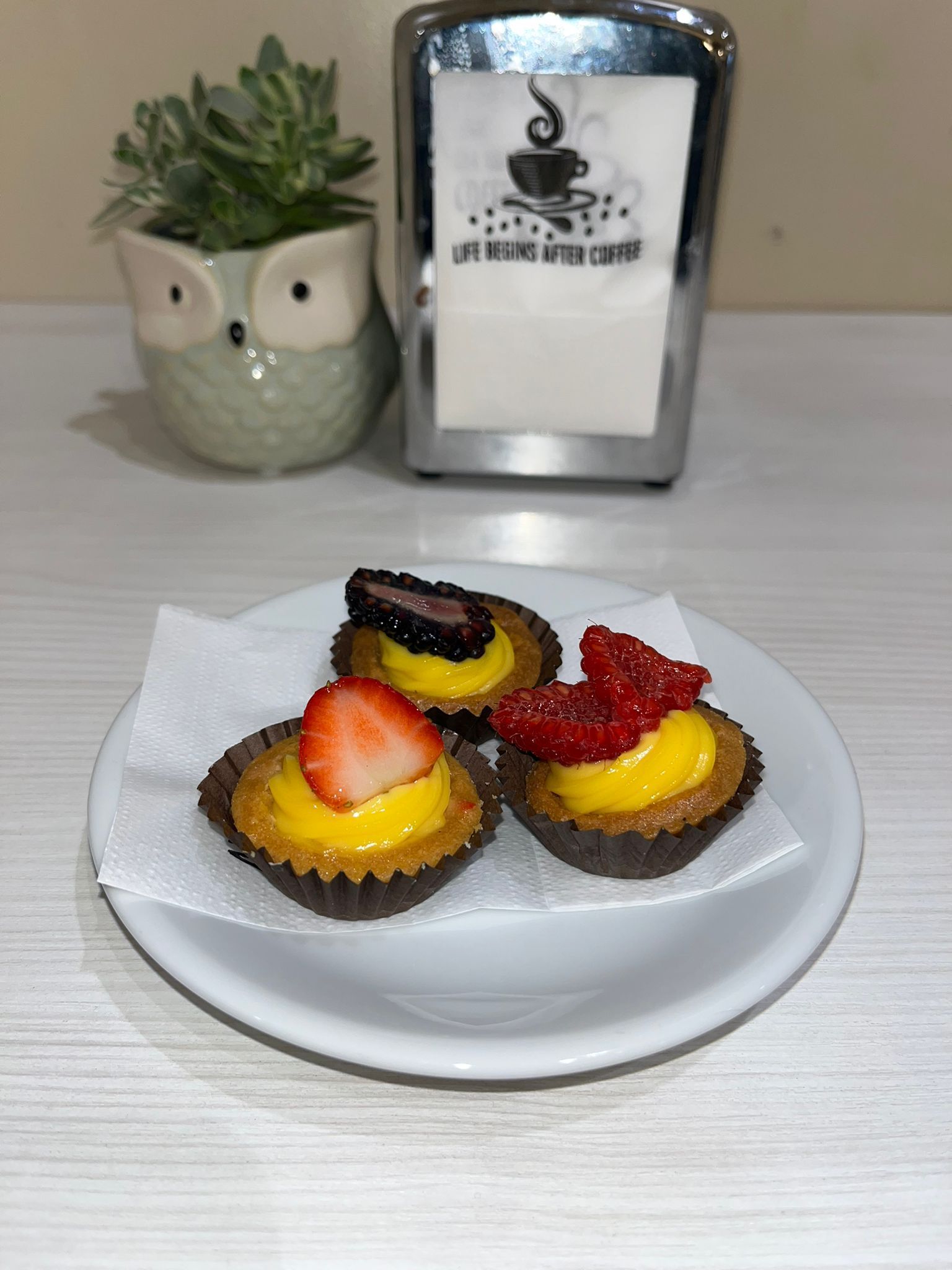 Tre tortine in miniatura su un piatto bianco, guarnite con frutta e crema gialla, vicino a un porta tovaglioli e a un vaso a forma di gufo.