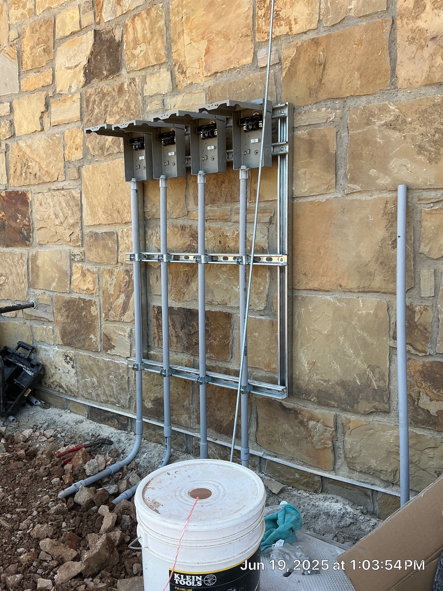 Electrical conduit and boxes mounted on a stone wall. A bucket and construction debris are in the foreground.