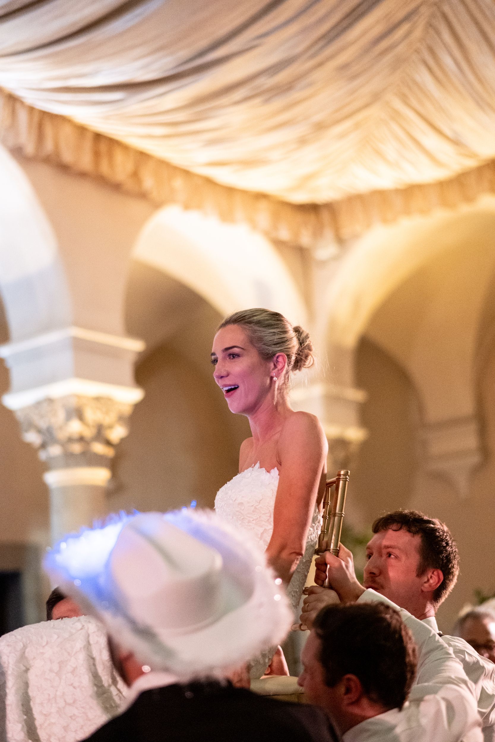A bride in a white dress is being lifted ona. chair on a man 's shoulders at a wedding reception.