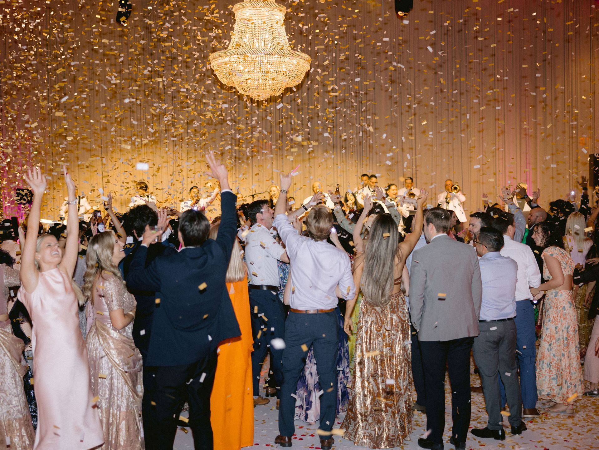 A large group of people are dancing at a wedding reception with confetti falling from the ceiling.