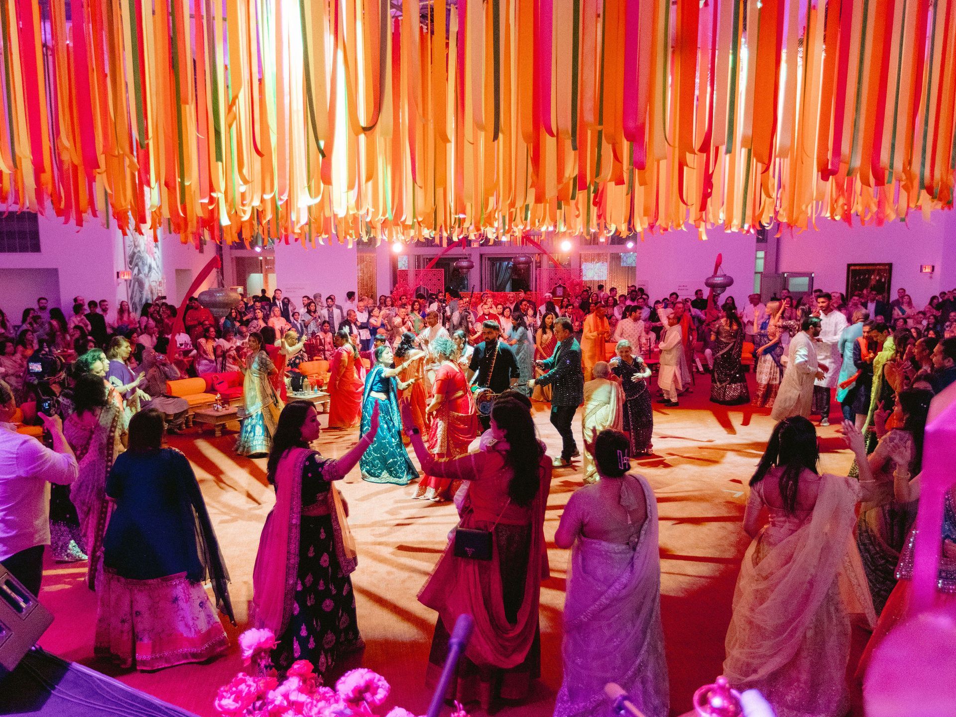 A large group of people are dancing in a room with colorful ribbons hanging from the ceiling.