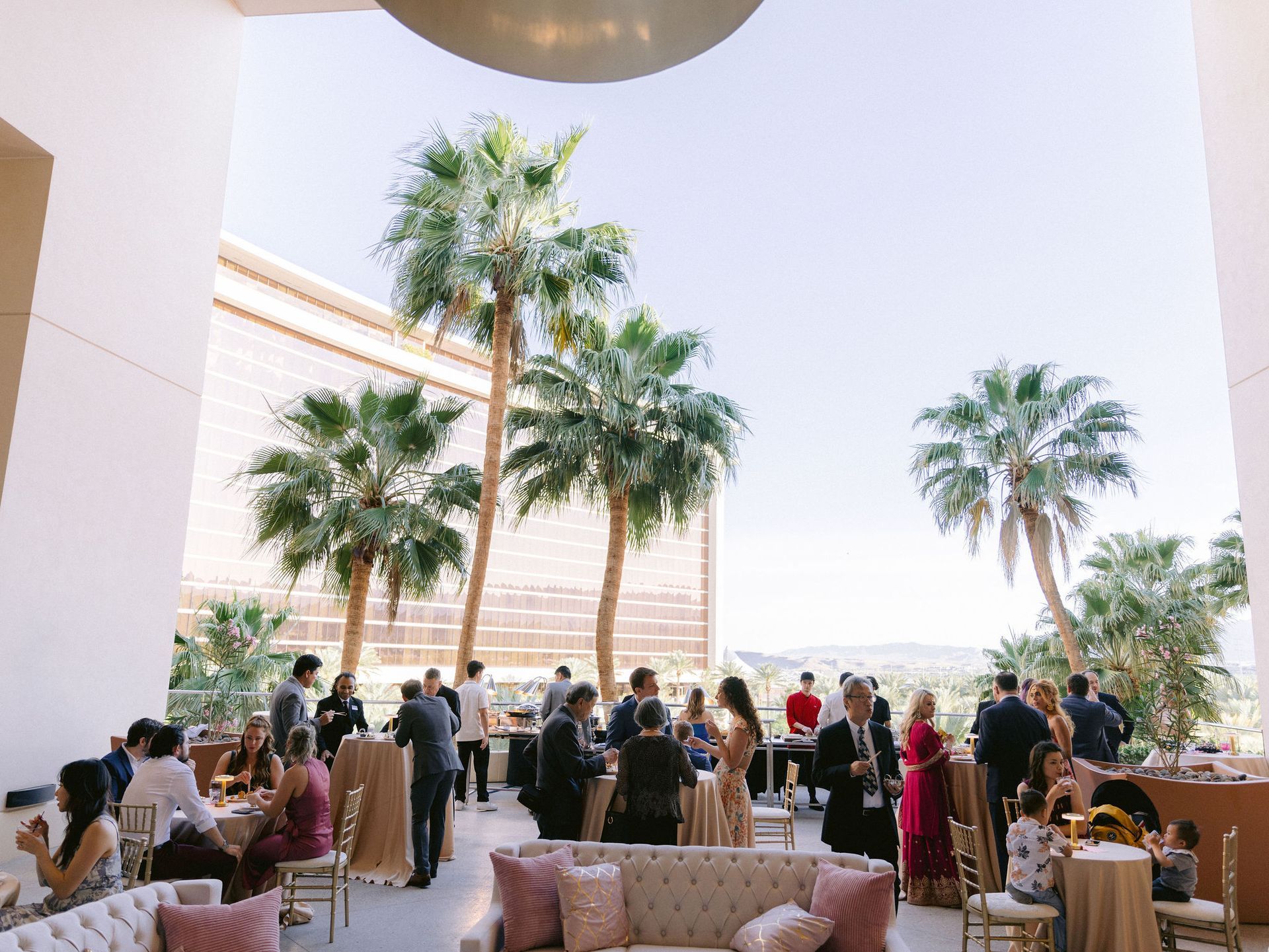 A group of people are standing around tables and chairs in a room with palm trees.