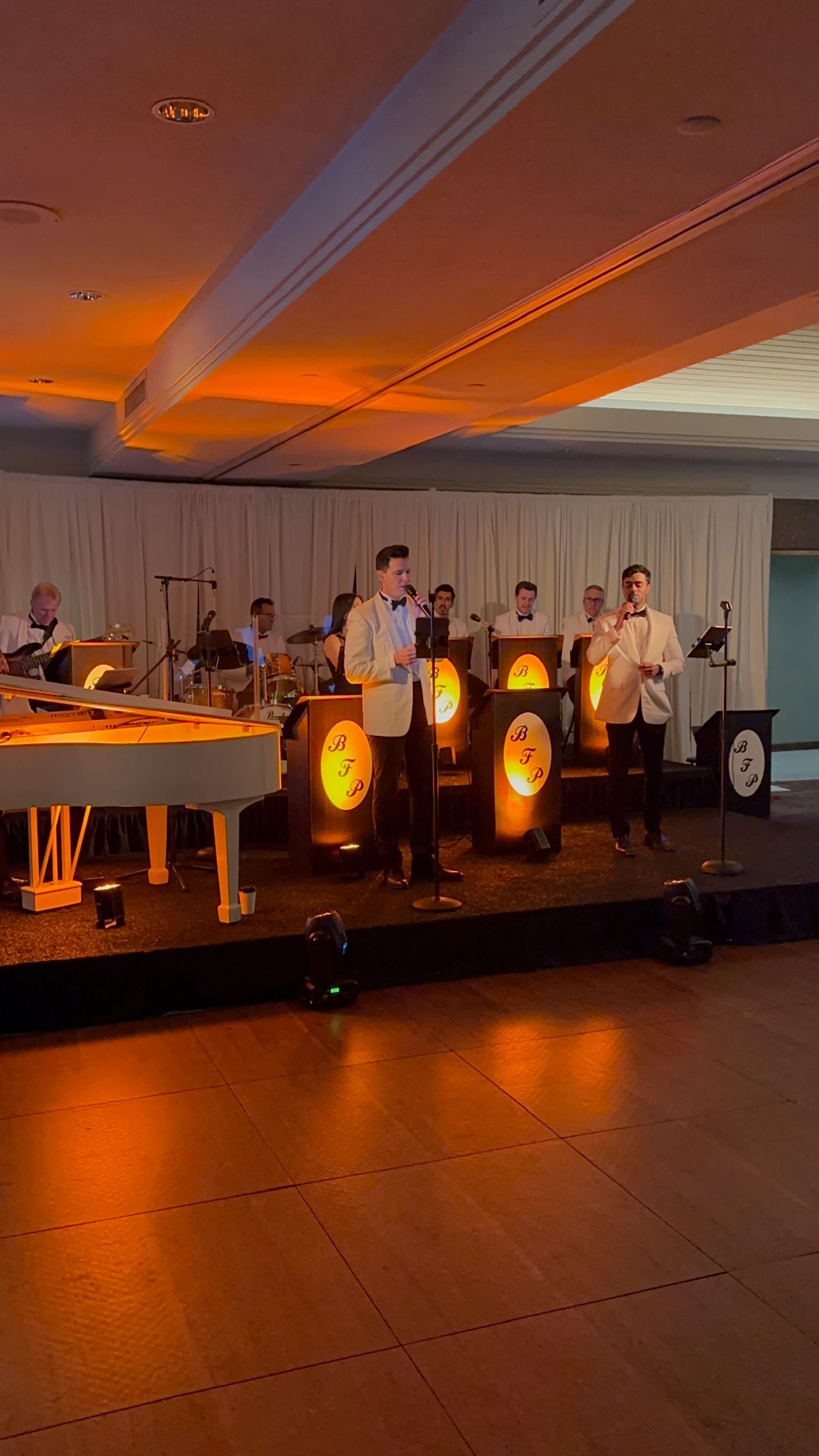 Band performing on a stage. Two singers stand in front, with a white piano and other musicians visible in the background, all lit with warm orange light.