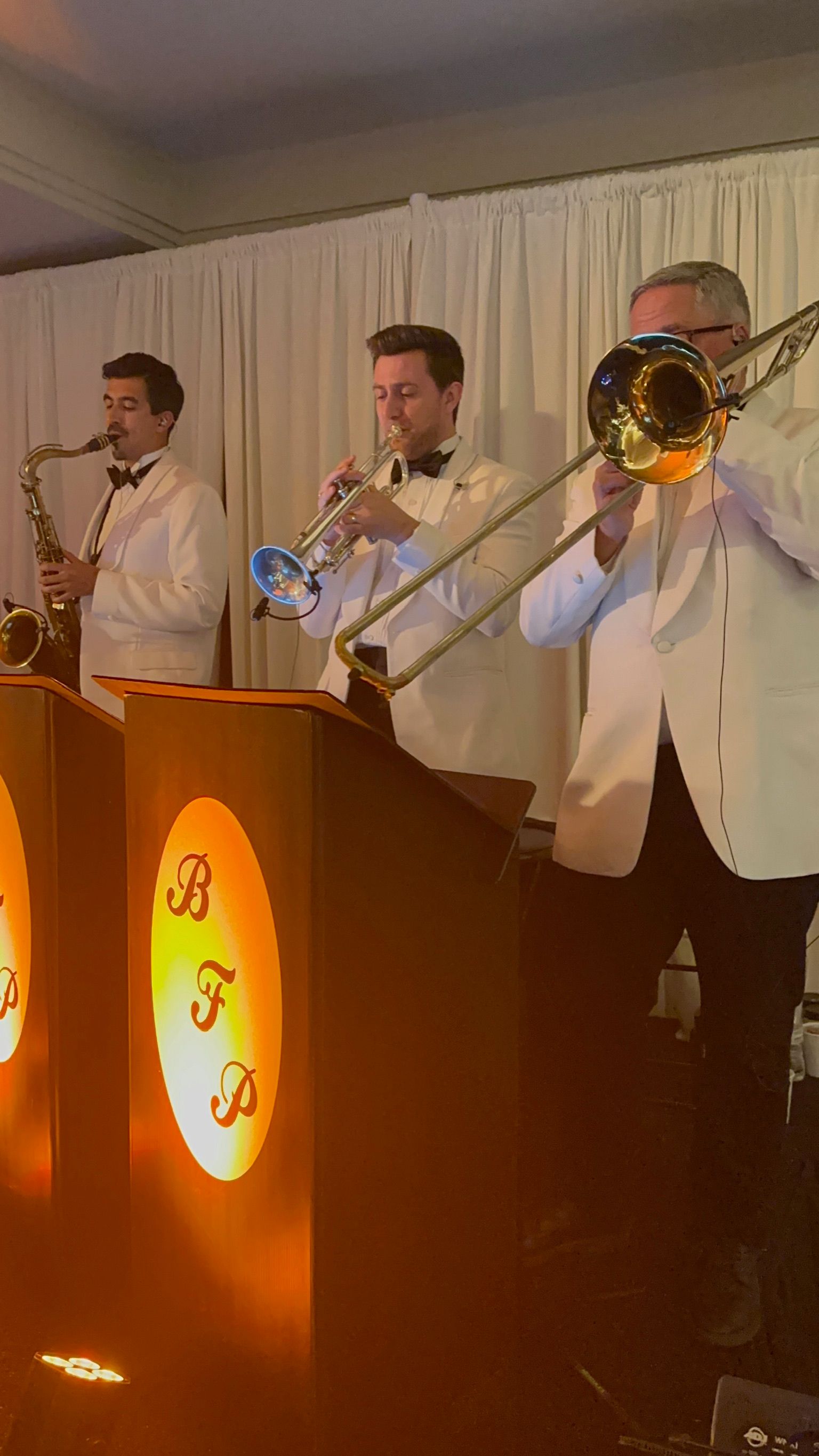 A jazz band in white suits and bow ties performs under warm lighting. Instruments include a saxophone, trumpet, and trombone.