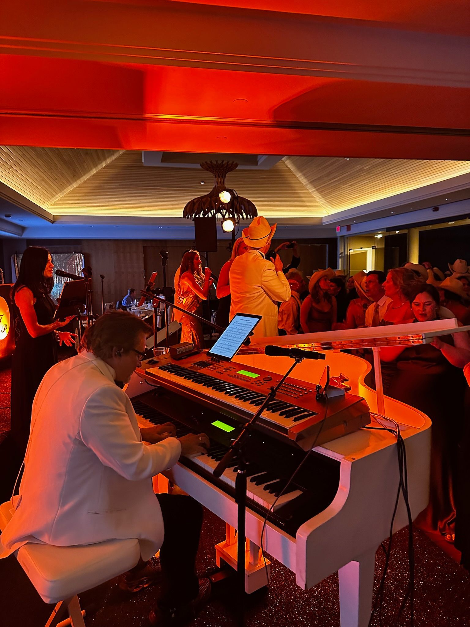 A band performs on a stage in a room lit with red lights. A man in a white suit plays a white piano. Other band members are visible behind him, and people are in the audience.