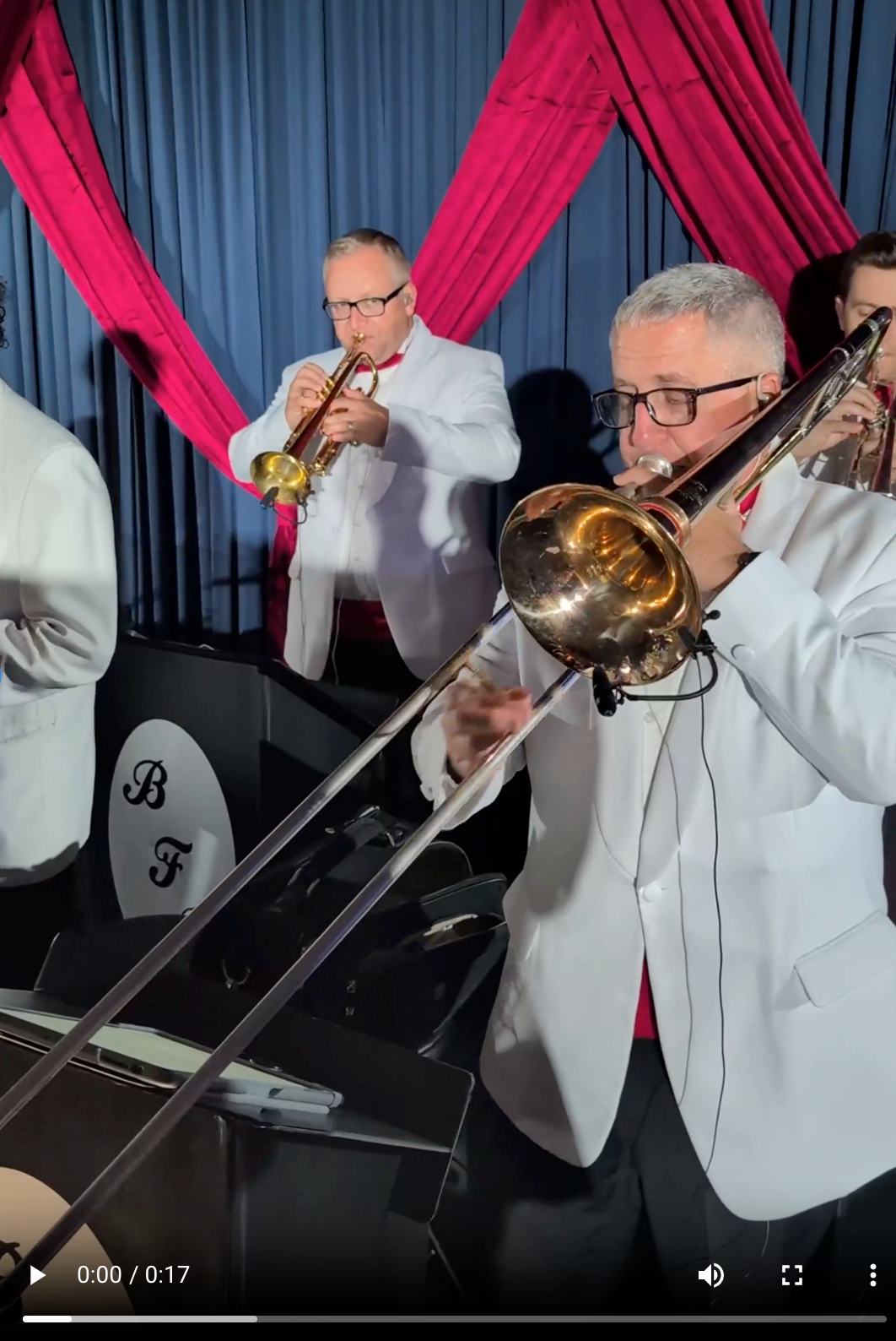 Band playing, two men in white suits with red bowties, trombonist in focus, trumpet player behind them, pink and blue backdrop.